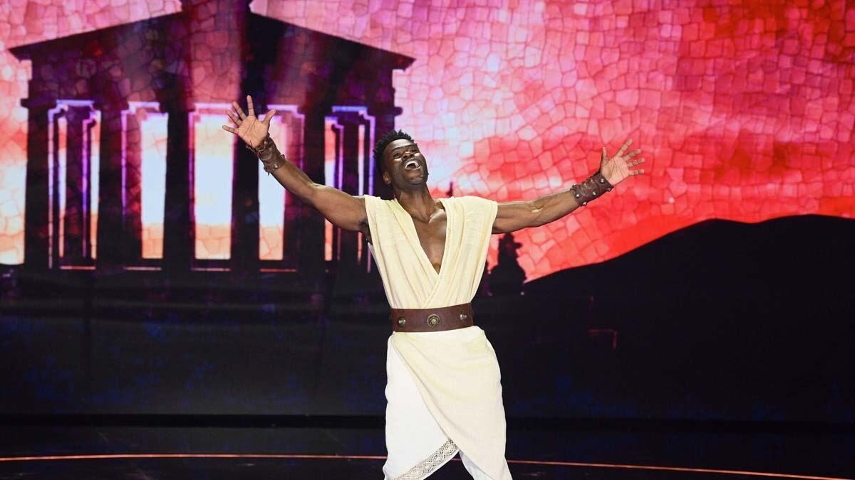 Hercules performer singing on stage with his arms out in front of a coliseum and red mosaic back drop 
