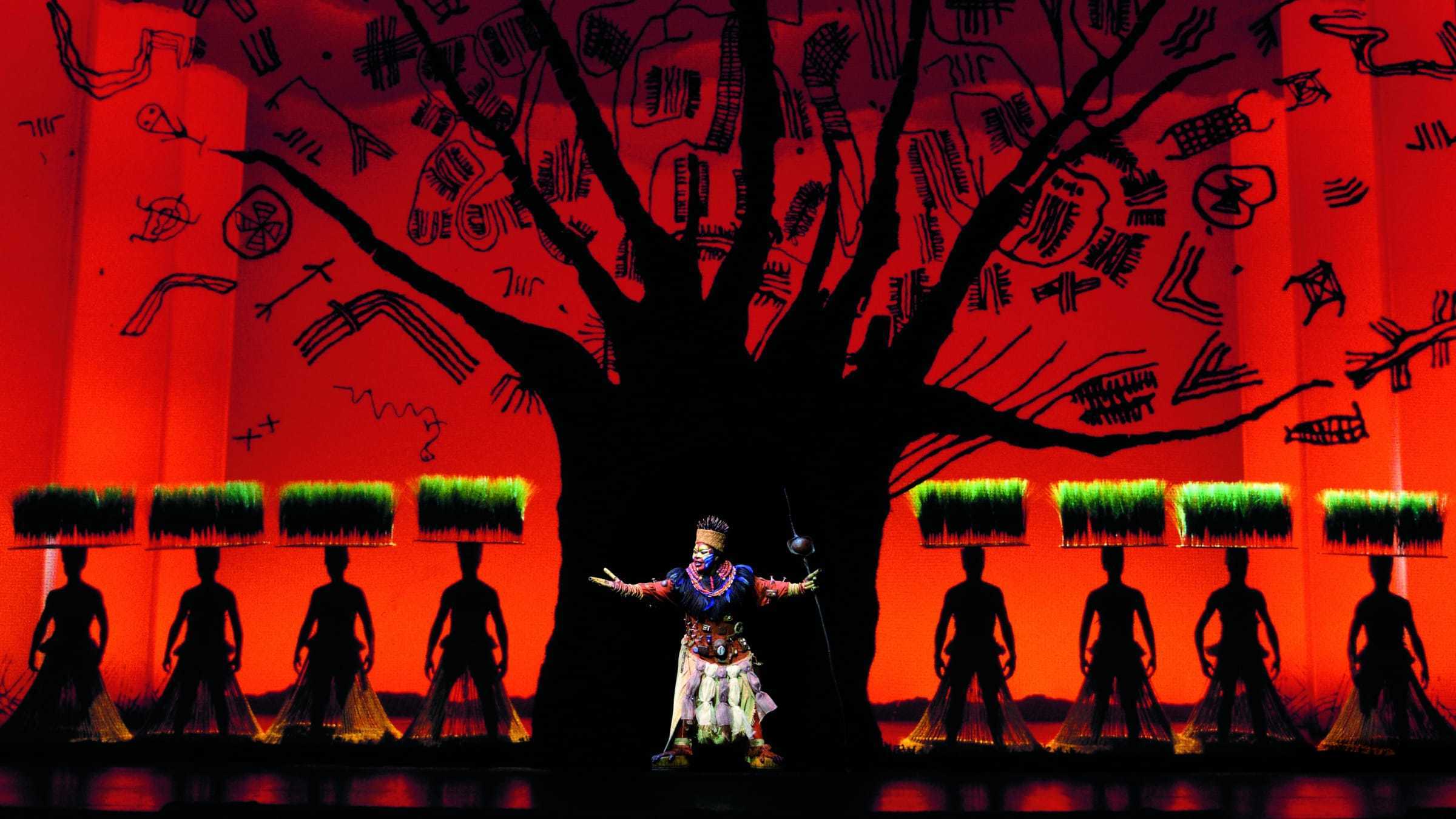 Rafiki character in costume on stage with cast members depicting grass in the background. 