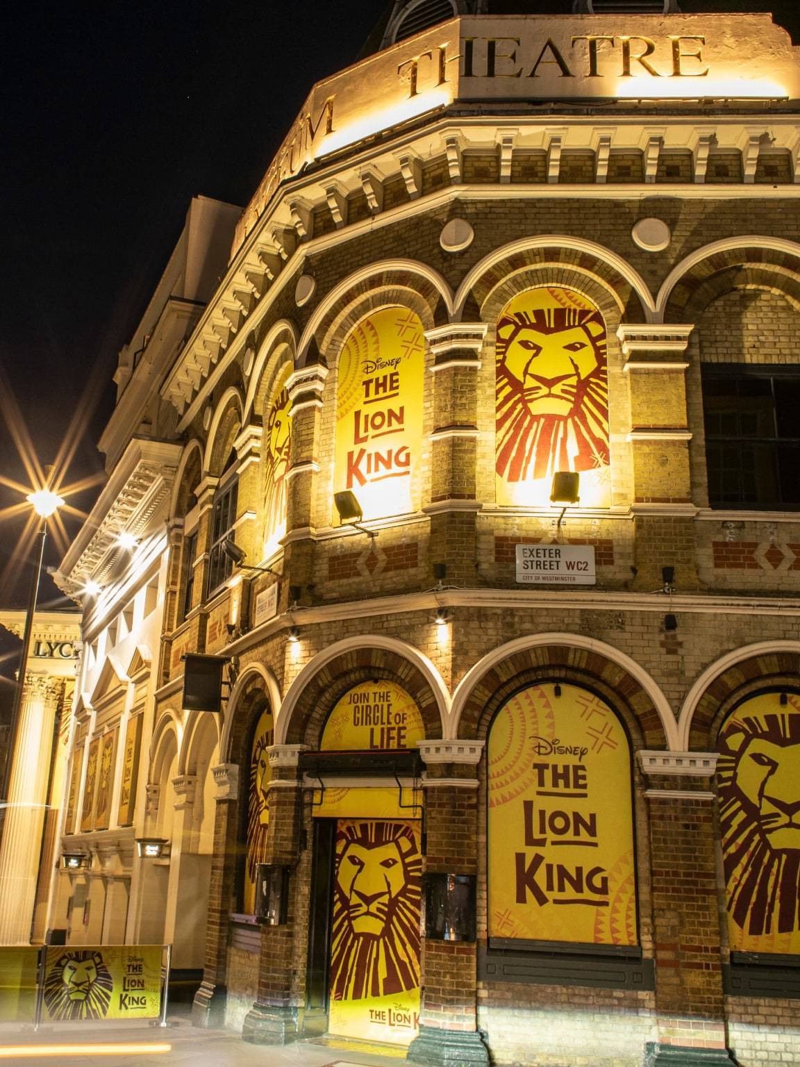 The exterior of the Lyceum Theatre at night with The Lion King posters lit up with lights. 