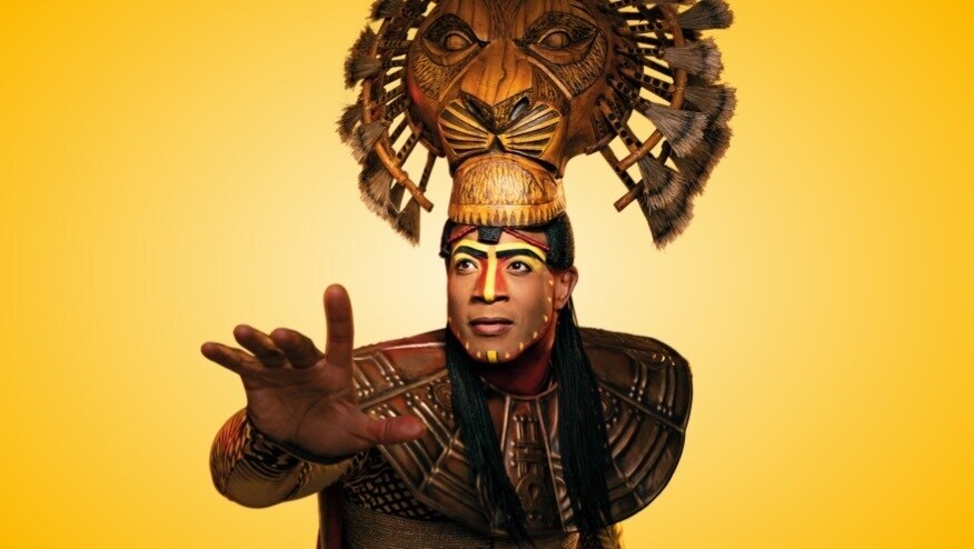 Yellow background with actor who plays Simba, in costume reaching his hand out 