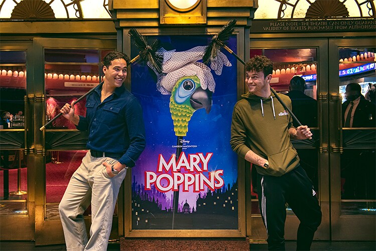 Louis Gaunt and Charlie Stemp smiling at each other in front of the Mary Poppins poster in the theatre's entrance.