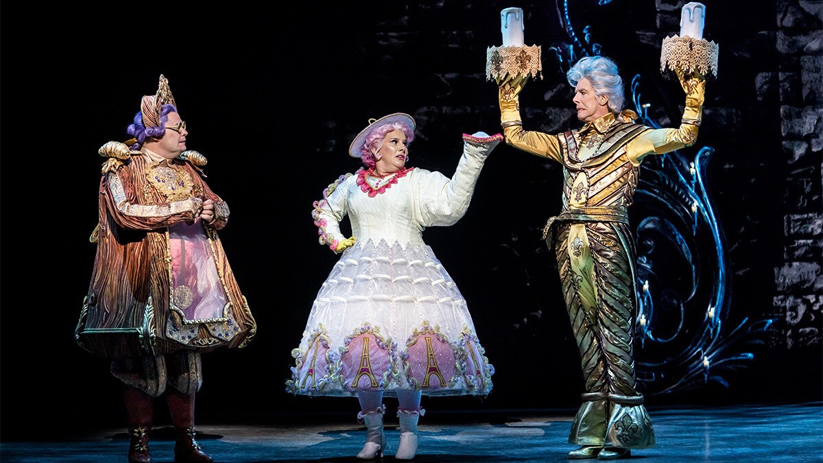 Actors playing Cogsworth, Mrs Potts and Lumiere looking at each other in confusion on the stage.