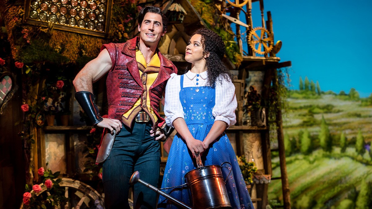 Gaston and Belle smiling in front of a little house on a village background.