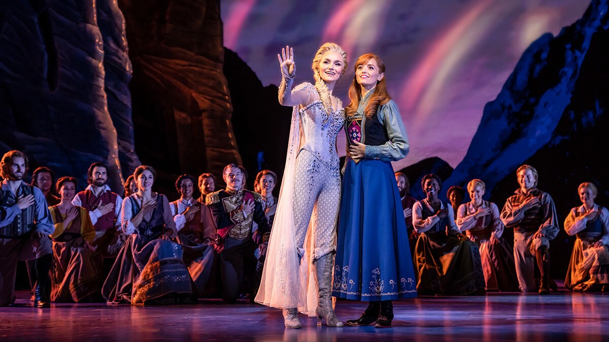 Elsa and Anna looking towards the audience happily with ensemble kneeling behind them with hand on their heart.