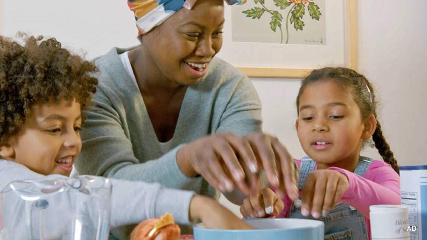 A woman with two young children, placing their hands into a mixing bowl